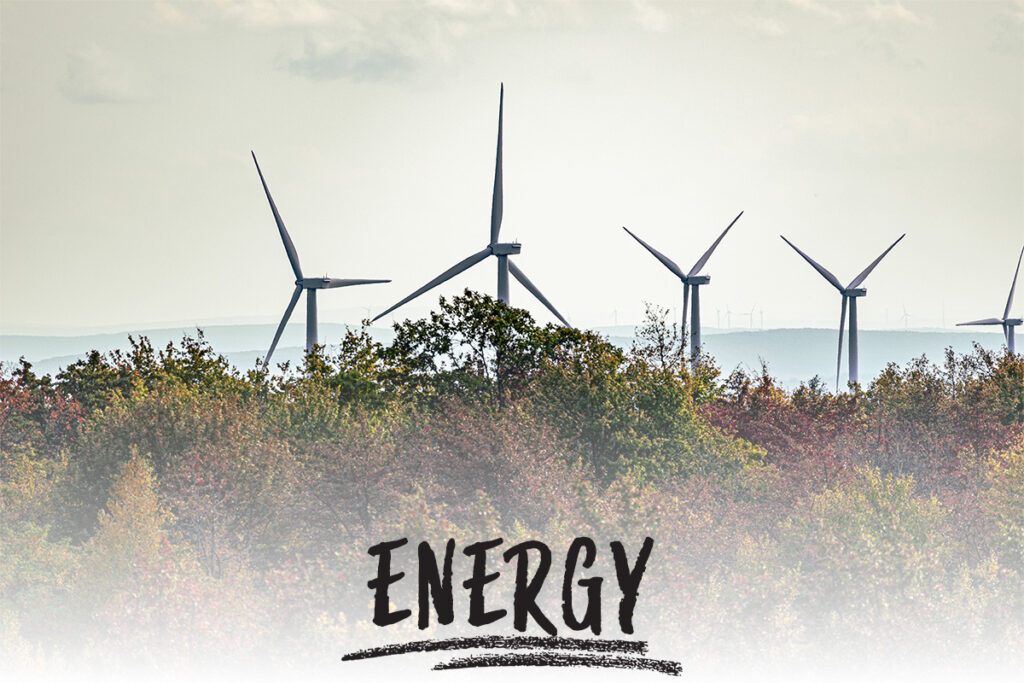 several windmills on top of a ridgeline with trees in the foreground and a ridgeline with more windmills in the background