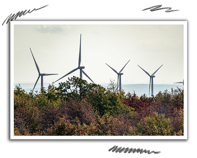 several windmills on top of a ridgeline with trees in the foreground and a ridgeline with more windmills in the background