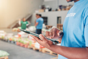 man in volunteer shirt in foreground entering information into a tablet while rows of food and volunteers stand in background