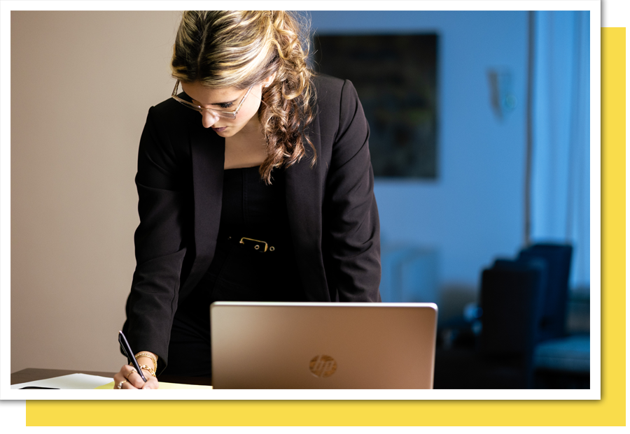 a woman standing and writing on a legal pad with a laptop in front of her